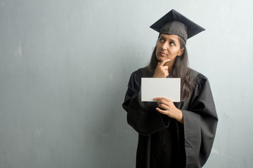 Young graduated indian woman against a wall thinking and looking up, confused about an idea, would be trying to find a solution. Holding a placard.