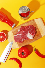 Raw beef meat on a wooden board and ingredients for cooking on a yellow background.