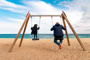 father and daugher having family playing on swing on the beach during autmn day in front of sea