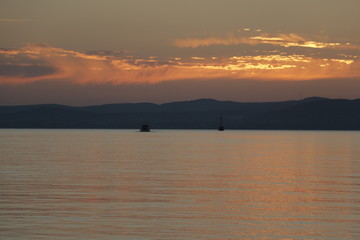 Two ships sailing on Lake Balaton. A warm, pleasant summer evening. A quiet end of the day. Beautiful sunset. Orange sky. Reflection of the sun on the surface of the water. Mountains in the background