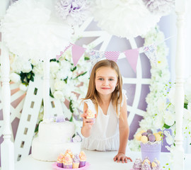 Obraz na płótnie Canvas Pretty little smiling girl standing at the white table with cakes in the beatiful decorated candy bar