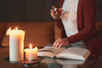Young woman sitting on the sofa at home and reading a thick book and drink tea. Cosy and relaxing atmosphere with candle light. Self Education Concept. Focus on foreground, blurred background. 