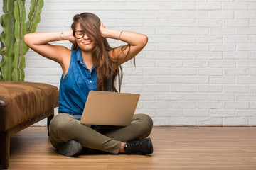 Portrait of young latin woman sitting on the floor covering ears with hands, angry and tired of hearing some sound. Holding a laptop.