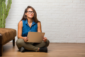 Portrait of young latin woman sitting on the floor looking up, thinking of something fun and having an idea, concept of imagination, happy and excited. Holding a laptop.