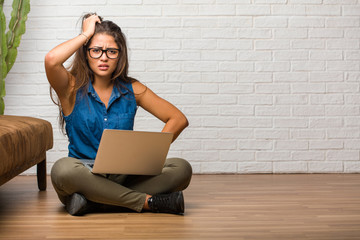 Portrait of young latin woman sitting on the floor worried and overwhelmed, forgetful, realize something, expression of shock at having made a mistake. Holding a laptop.