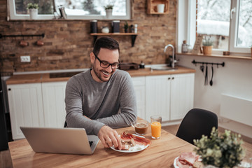 Handsome casual man having breakfast and using laptop in modern rustic kitchen.