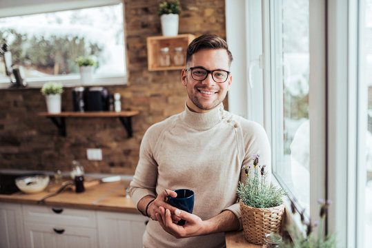 Portrait of smiling millenial man drinking tea near the window at cozy home on winter morning.