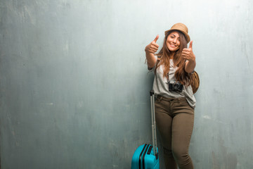 Portrait of young traveler latin woman against a wall cheerful and excited, smiling and raising her thumb up, concept of success and approval, ok gesture. Holding a blue suitcase.