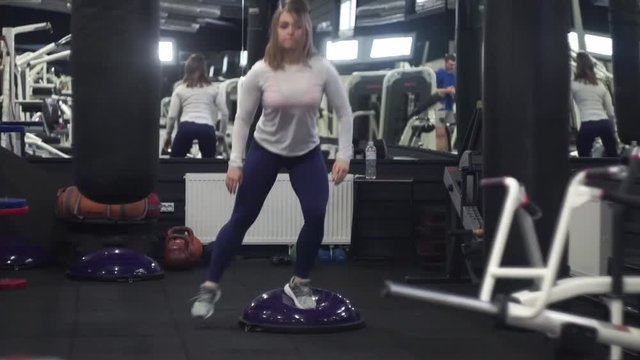 Female jumping on bosu doing cardio aerobics workout in gym. She is alone and concentrated on achieving result. Long shot