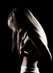 Beautiful slim topless blonde girl with hair falling on her face, stands sideways with a her hand covering her big boobs. Black background. Artistic noir silhouette photo