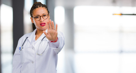 Portrait of a young black doctor woman serious and determined, putting hand in front, stop gesture,...