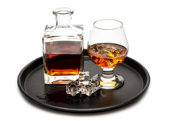 A glass of whiskey with ice or brandy and a square carafe on a black dish isolated against a white...