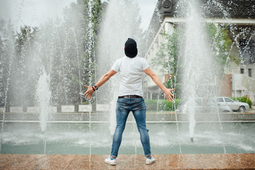 Picture of young man in white tshirt and blue jeans standing in front of fountain. Splashing upward cool transparent water.