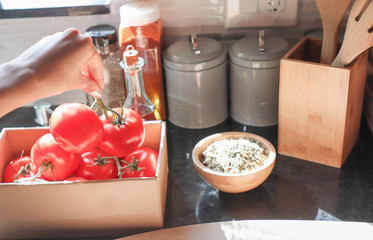 Female hand takes out of the box on the twig a tomatoes. A box with tomatoes is on the kitchen worktop. olive oil and spices