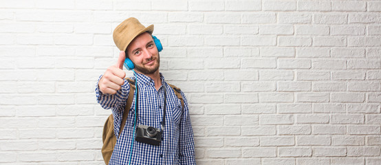Young traveler man wearing backpack and a vintage camera cheerful and excited, smiling and raising her thumb up, concept of success and approval, ok gesture. Listening to music with headphones.