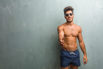 Young athletic man wearing a swimsuit against a grunge wall reaching out to greet someone or gesturing to help, happy and excited