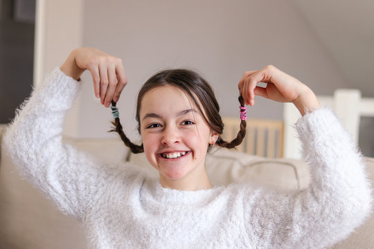 portrait of stylish foolish attractive smiling preteen girl holding up her pigtails and making faces sitting on sofa at home. Having fun at April fools day. Playful childhood emotions.