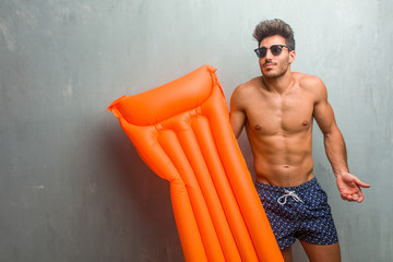 Young athletic man wearing a swimsuit against a grunge wall doubting and shrugging shoulders, concept of indecision and insecurity, uncertain about something - obrazy, fototapety, plakaty