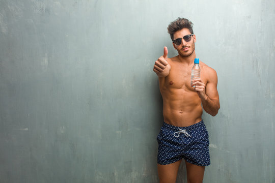 Young athletic man wearing a swimsuit against a grunge wall cheerful and excited, smiling and raising her thumb up, concept of success and approval, ok gesture