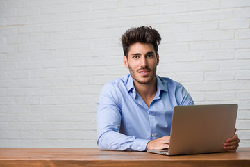 Young business man sitting and working on a laptop with hands on hips, standing, relaxed and smiling, very positive and cheerful