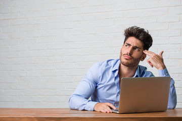 Young business man sitting and working on a laptop making a suicide gesture, feeling sad and scared forming a gun with fingers