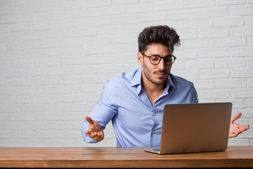 Young business man sitting and working on a laptop doubting and shrugging shoulders, concept of indecision and insecurity, uncertain about something - obrazy, fototapety, plakaty