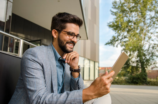 Elegant man in suit sitting on bench in front of office building and looking at tablet 