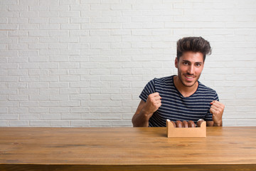 Young handsome and natural man sitting on a table very happy and excited, raising arms, celebrating a victory or success, winning the lottery. Eating chocolate doughnuts.