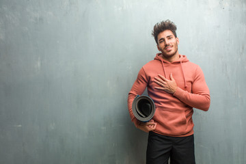 Young fitness man against a grunge wall doing a romantic gesture, in love with someone or showing affection for some friend. Holding a blue mat for practicing yoga.