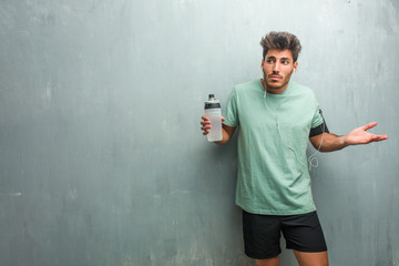 Young fitness man against a grunge wall doubting and shrugging shoulders, concept of indecision and...
