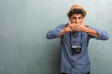 Young handsome traveler man wearing a straw hat, a backpack and a photo camera covering mouth,...