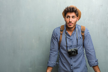 Young handsome traveler man wearing a straw hat, a backpack and a photo camera doubting and shrugging shoulders, concept of indecision and insecurity, uncertain about something - obrazy, fototapety, plakaty