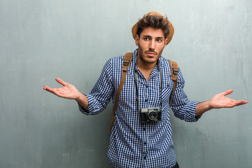 Young handsome traveler man wearing a straw hat, a backpack and a photo camera doubting and shrugging shoulders, concept of indecision and insecurity, uncertain about something - obrazy, fototapety, plakaty