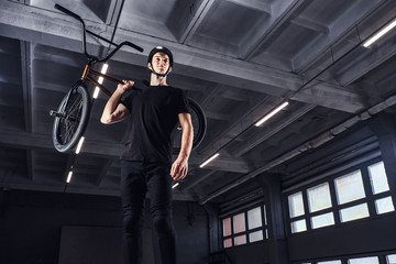 Full body portrait of a BMX rider holding his bike on shoulder and looking away in a skatepark indoors