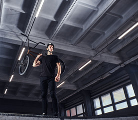 Full body portrait of a BMX rider holding his bike on shoulder and looking away in a skatepark indoors