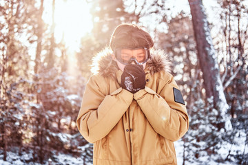 Young snowboarder dressed in warm clothes and goggles, warms his hands standing in the snowy forest...