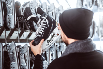 A young guy wearing warm clothes standing next to a rack and choosing a pair of skates in a skate hire inside.