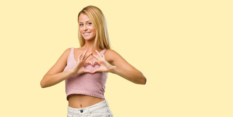 Portrait of young pretty blonde woman making a heart with hands, expressing the concept of love and friendship, happy and smiling