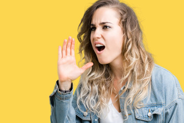 Beautiful young blonde woman wearing denim jacket over isolated background shouting and screaming loud to side with hand on mouth. Communication concept.