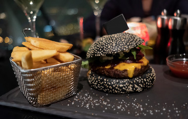 Tasty black burger, French fries in basket and ketchup served on black cutting board. Copy-space flag stuck into bun for your individual text