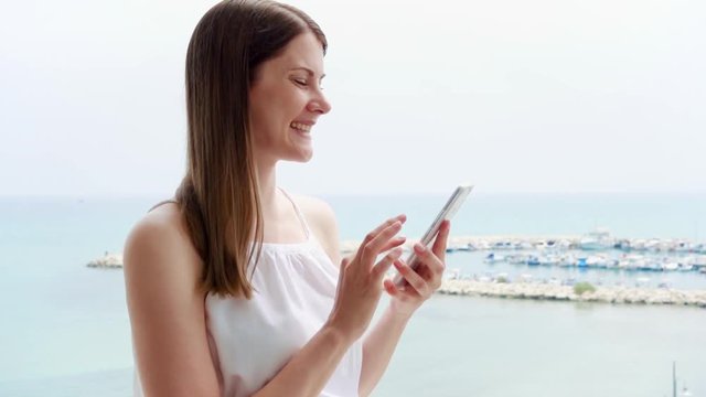 Young woman in white dress using mobile phone on balcony. Female on vacation enjoying sea view from terrace in morning. Smiling girl browsing on cellphone in slow motion