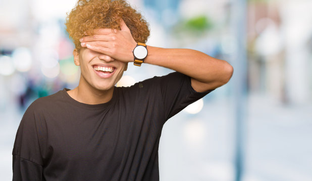 Young handsome man with afro hair wearing black t-shirt smiling and laughing with hand on face covering eyes for surprise. Blind concept.