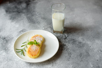 Tasty fresh cottage cheese pancakes on a white plate with a glass of milk on a concrete background. Healthy and diet breakfast
