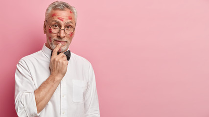 Curious pensioner has thick grey beard keeps finger on lips, looks with interest, dressed in formal white shirt, recieves many kisses, has traces on face, models on pink wall with empty space