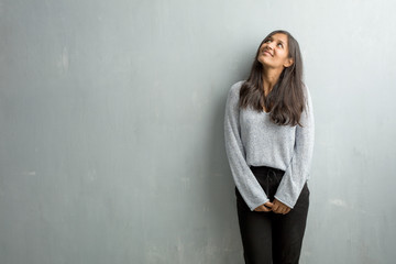 Young indian woman against a grunge wall looking up, thinking of something fun and having an idea, concept of imagination, happy and excited