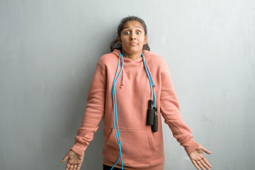Young sporty indian woman against a wall doubting and shrugging shoulders, concept of indecision and insecurity, uncertain about something. Holding a jump rope. - obrazy, fototapety, plakaty