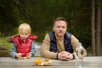 Little boy and his father is eating snacks during picnic on coast of mountain river in Switzerland.