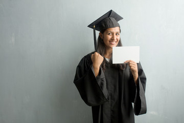 Young graduated indian woman against a wall very happy and excited, raising arms, celebrating a victory or success, winning the lottery. Holding a placard.