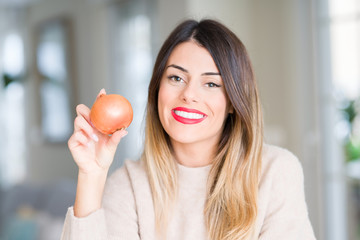 Young beautiful woman holding fresh onion at home with a happy face standing and smiling with a confident smile showing teeth