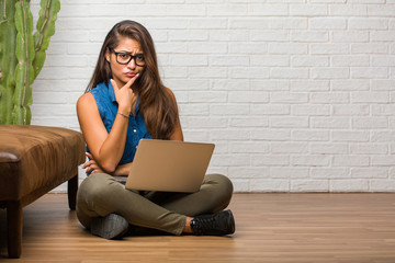 Portrait of young latin woman sitting on the floor doubting and confused, thinking of an idea or worried about something. Holding a laptop.